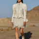 A model walks confidently on a stone path in the desert during the Louis Vuitton Cruise 2026 show. She wears a structured, ivory leather jacket with a flared, sheer chiffon skirt and matching ankle boots. Her look is completed with sleek, futuristic sunglasses, framed by a clear blue sky and sunlit desert mountains in the background.